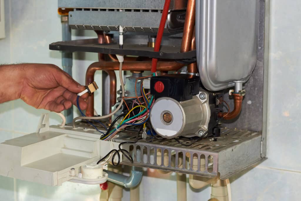 Réparation chaudière gaz : l'entretien expert de votre appareil Technicien réparant une chaudière à gaz murale, avec ses composants internes exposés: tuyaux cuivre, câbles et moteur.