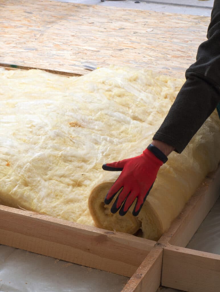 Isolation de plancher : Pose de laine minérale facile Main gantée rouge déroulant une couverture de laine minérale jaune pour isoler un plancher en bois avec des chevrons.