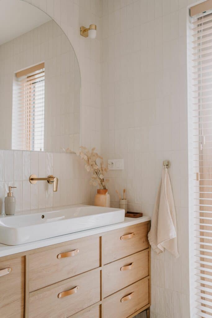 Salle de bain moderne: meuble bois, vasque blanche, touches dorées Salle de bain moderne: vasque blanche, meuble en bois clair, robinet mural doré, miroir arqué. Mur carrelé blanc, stores clairs, détails naturels.